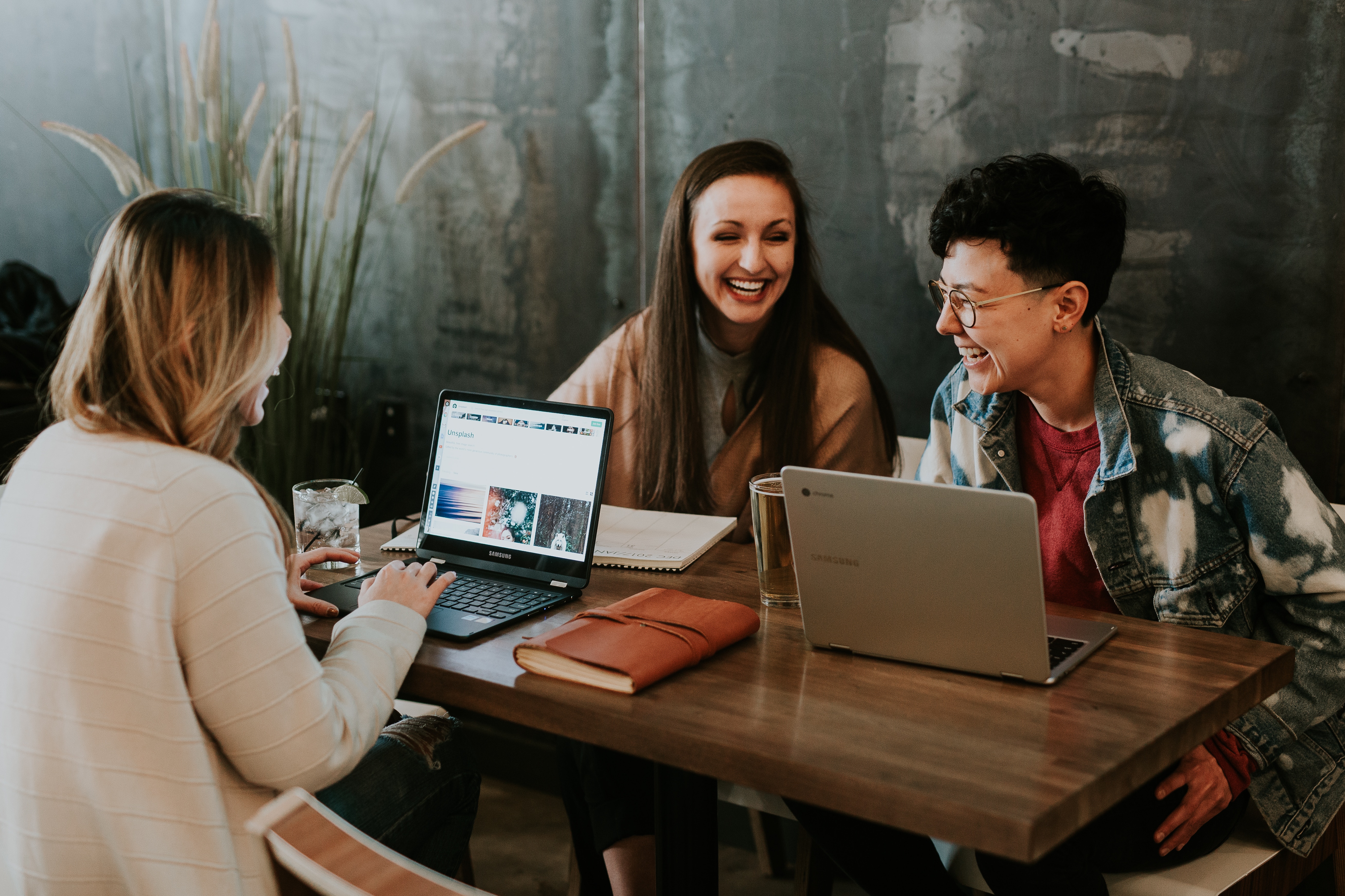 Three twenty-something people sit at a table with laptops and notebooks, laughing.